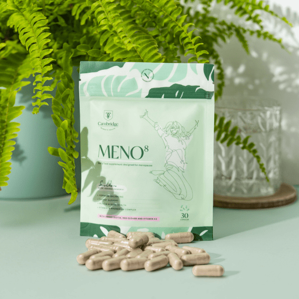 A packet of Meno8 with the about 30 pills in front. The pills are light brown in colour and cylinder shaped. Behind is a fern house potted plant and a glass on a coaster.